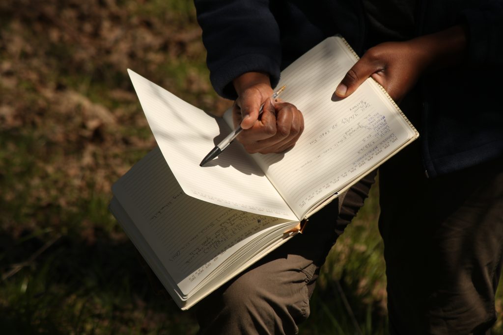 A close-up view of the hands of a researcher who is writing field notes down in his notebook. He is using a black pen.