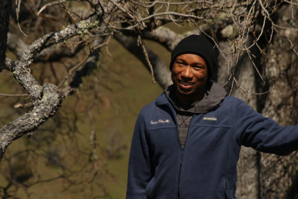 A profile photo of a young man standing in front of an oak tree, with his arm outstretched and smiling at the camera. He is wearing a beanie and a zip-up sweater that says "Smith Fellows" on it.
