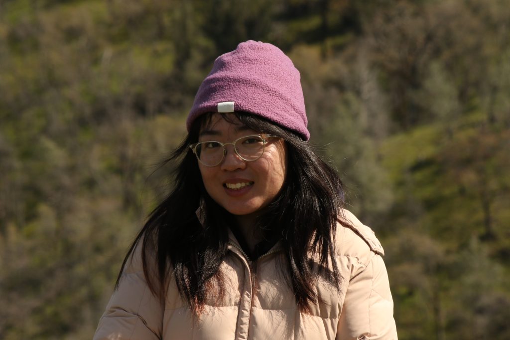 Profile photo of a young woman wearing glasses, a beige puffy jacket, and a pink beanie. She is smiling at the camera. In the background is an oak woodland habitat.