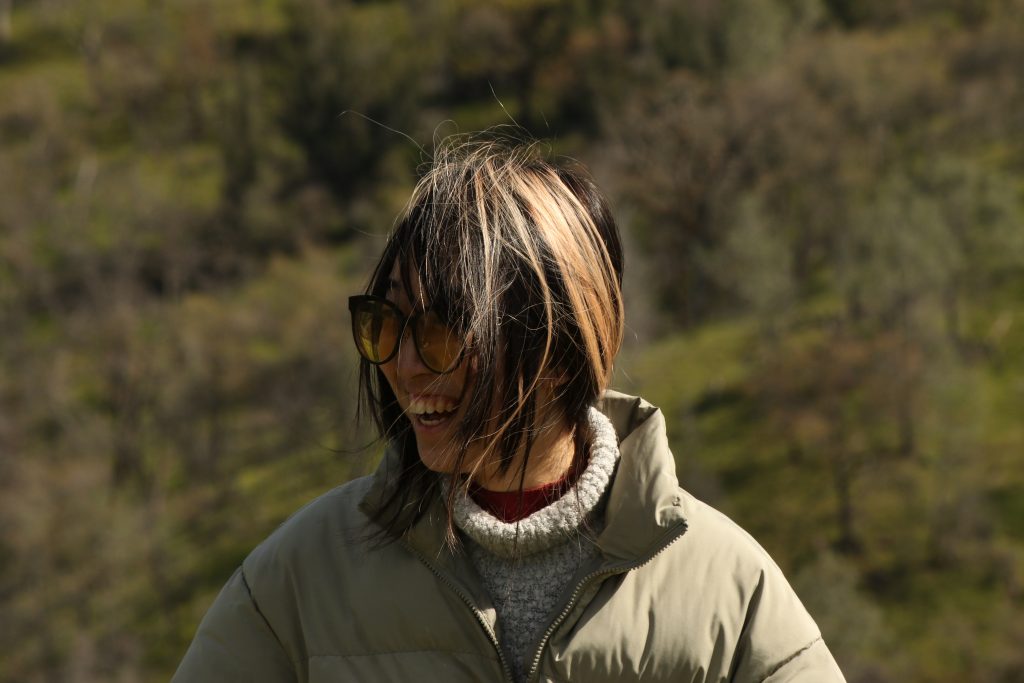 Profile photo of a young woman wearing glasses, a beige puffy jacket, and blonde highlighted black hair. She is smiling, looking sideways away from the camera. In the background is an oak woodland habitat.